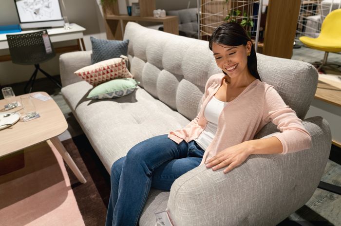 A woman sits on a grey couch at a furniture store, smiling and resting her hand on the armrest.
